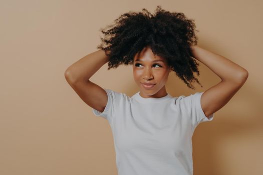 Feeling gorgeous. Portrait of young attractive sensual african woman touching with hands her curly hair and looking aside with flirty look, dressed in white basic tshirt, isolated on beige background