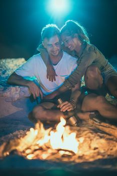 A happy family is sitting on the sandy seashore by the fire in the evening. They are making fun and drinking beer.