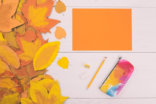 Empty orange paper and school supplies on wooden table with painted leaves.Toned image.