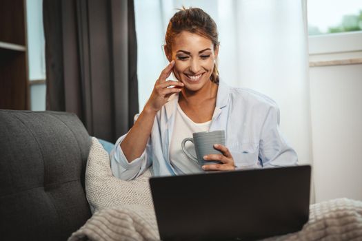 Beautiful young woman sitting on the sofa, enjoying a cup of coffee and using her laptop at home.