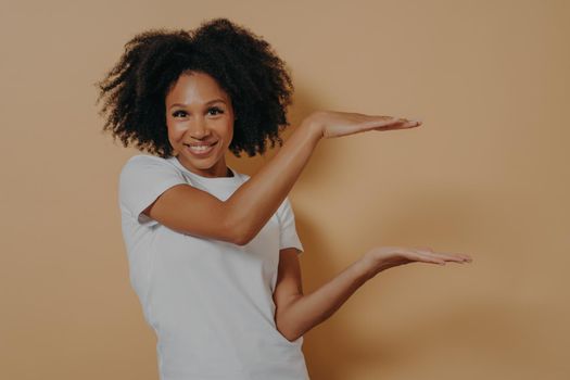 Young cheerful dark skinned showing large object, shaping box with both hands and smiling at camera while posing isolated over beige wall with copy space for text. Advertisement concept