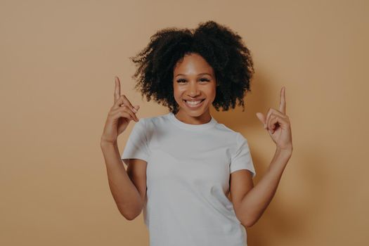 Young overjoyed excited dark skinned woman with curly hair wearing white tshirt pointing up with forefingers and showing blank copy space for advertising, isolated on beige studio background