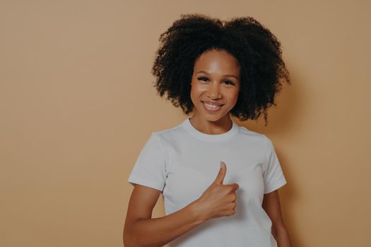 Cheerful positive young african woman showing success gesture, thumb up with hand and smiling at camera with positive face expression, saying well done, isolated on beige studio background