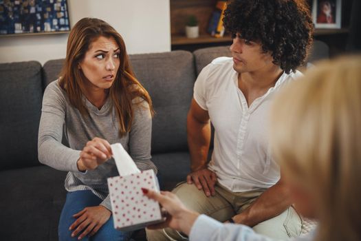 Shot of a female psychotherapist offering tissues to her young couple patients during a counseling session on a sofa inside of a living room.