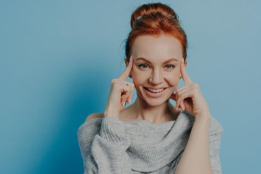 Headshot of young positive red haired woman with fingers on temples smiling at camera while standing isolated on blue background, thinking and trying to focus on task, dressed in knitted sweater