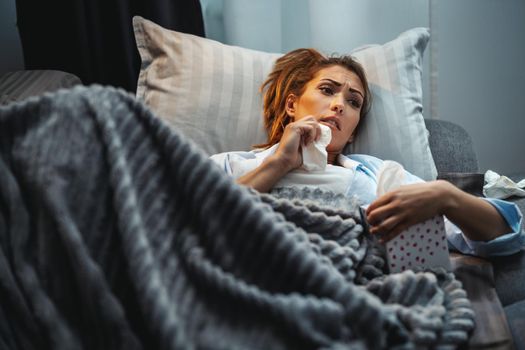 Young woman is lying sick at home couch with paper handkerchief in her hand for blowing nose.