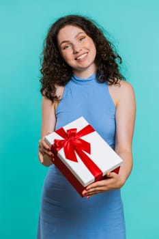 Positive smiling young woman presenting birthday green gift box stretches out hands, offer wrapped present career bonus, celebrating party sale. Girl isolated alone on blue studio background. Vertical