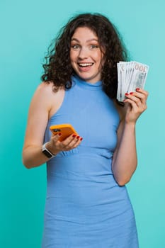 Young woman looking smartphone display sincerely rejoicing win, receiving money dollar cash banknotes, celebrating success lottery luck. Adult girl isolated alone on blue studio background. Vertical