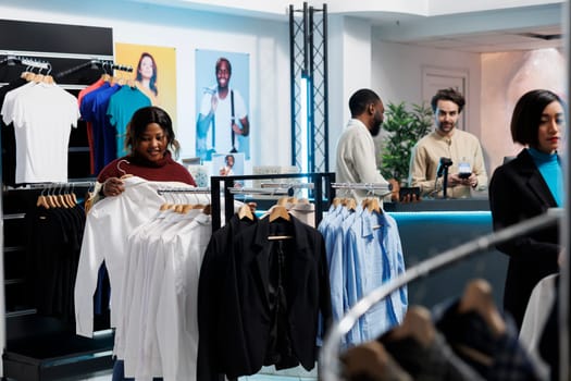 African american woman browsing through rack of formal wear while choosing outfit in clothing store. Boutique department customer holding shirt on hanger, examining style and size