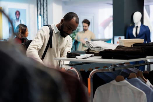 African american man browsing rack with casual clothes while shopping in mall fashion department. Boutique store customer examining menswear apparel while choosing trendy outfit