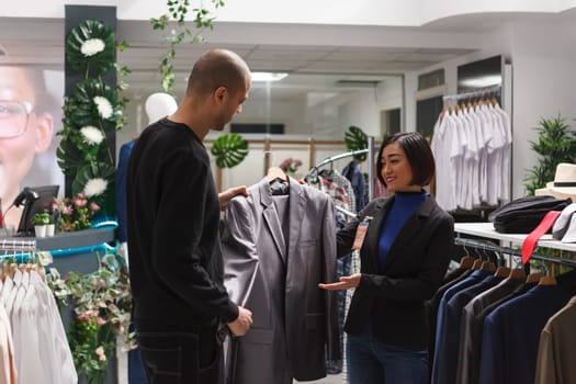 Shopping mall boutique smiling asian woman seller assisting arab man client in selecting jacket. Clothing store worker holding formal apparel hanger, showing outfit to customer, giving fashion advice