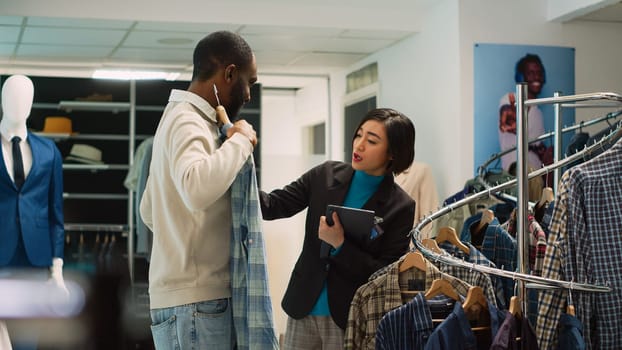 Diverse people examining clothes on racks to do shopping, woman working as store assistant in shopping center to help man choose shirts. Client looking at fashion merchandise. Handheld shot.