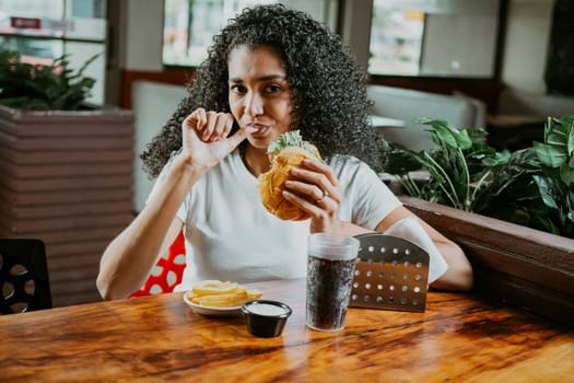 Latin woman sucking her fingers holding a hamburger in a restaurant. Portrait of an afro girl enjoying hamburger in a restaurant