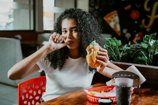 Portrait of an afro girl enjoying hamburger in a restaurant. Latin woman sucking her fingers holding a hamburger in a restaurant