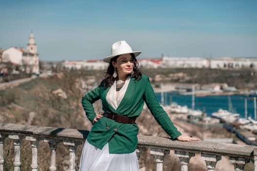 Woman walks around the city, lifestyle. Happy woman in a green jacket, white skirt and hat is sitting on a white fence with balusters overlooking the sea bay and the city