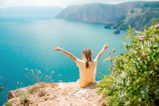 Happy girl perched atop a high rock above the sea, wearing a yellow jumpsuit and braided hair, signifying the concept of summer vacation at the beach