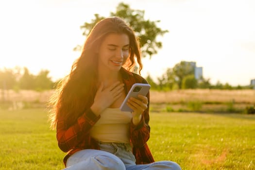 Excited young girl looks into the mobile phone sitting on the grass in the park at sunlight.