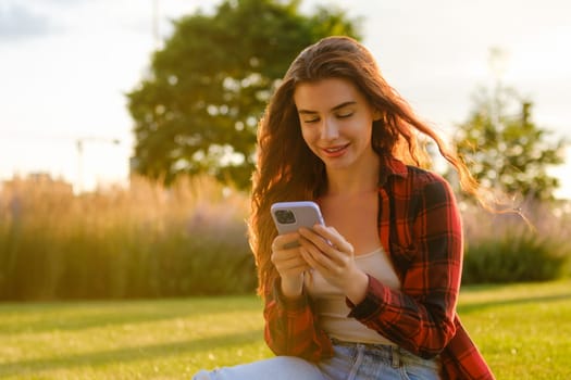 The happy millennial girl checking social media holding a smartphone sitting on the grass in the park at sunset.