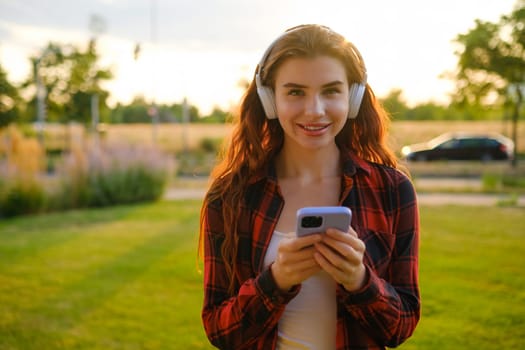 Happy young woman wearing wireless headphones for listening to the music via mobile phone in the park at sunset.