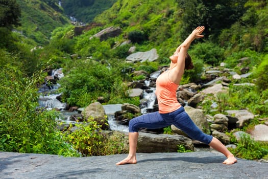 Yoga outdoors - sporty fit woman doing Ashtanga Vinyasa Yoga asana Virabhadrasana 1 Warrior pose posture at waterfall in HImalayas mountains