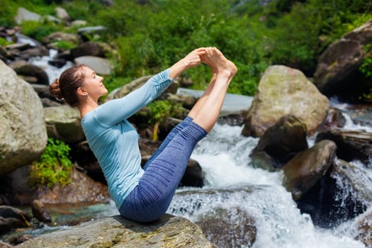 Yoga exercise outdoors - woman doing Ashtanga Vinyasa Yoga balance asana Ubhaya padangusthasana Big Double Toe Yoga Pose at tropical waterfall in Himalayas in India