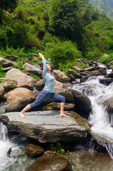 Yoga outdoors - sporty fit woman doing Ashtanga Vinyasa Yoga asana Virabhadrasana 1 Warrior pose posture at waterfall in HImalayas mountains