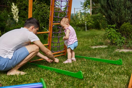 Dad with little child girl daughter collects wooden home sports complex outdoor. assembly process