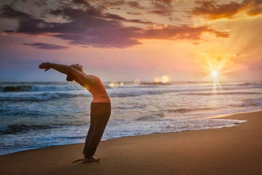 Young sporty fit woman doing yoga Sun salutation Surya Namaskar pose Hasta Uttanasana on tropical beach on sunset. With lens flare