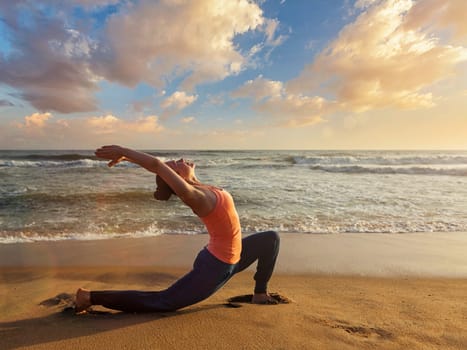 Yoga outdoors - sporty fit woman practices yoga Anjaneyasana - low crescent lunge pose outdoors at beach on sunset