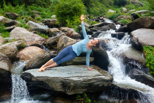 Yoga outdoors - beautiful sporty fit woman doing yoga asana Vasisthasana - side plank pose at tropical waterfall