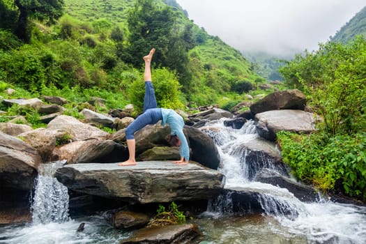 Yoga outdooors - woman doing yoga asana eka pada urdva dhanurasana Upward Bow Pose back benkd outdoors at waterfall in Himalayas