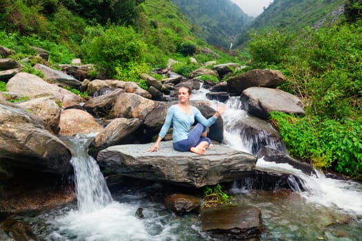 Yoga exercise outdoors - woman doing Ardha matsyendrasanaasana asana - half spinal twist pose at tropical waterfall in Himalayas in India
