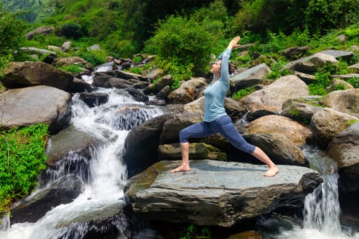 Yoga outdoors - sporty fit woman doing Ashtanga Vinyasa Yoga asana Virabhadrasana 1 Warrior pose posture at waterfall in HImalayas mountains