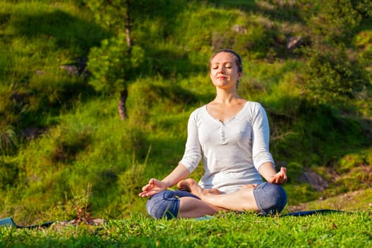 Young sporty fit woman doing yoga outdoors - meditating and relaxing in Padmasana Lotus Pose) with chin mudra on green grass in forest