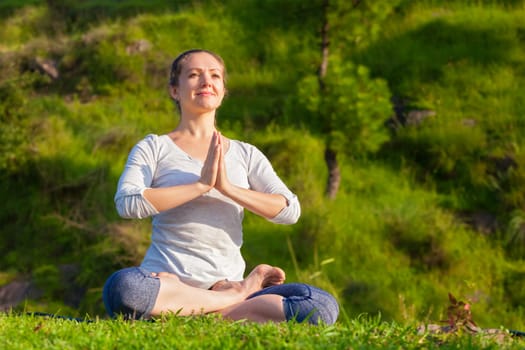 Meditation and relaxation yoga outdoors - young woman meditating and relaxing in Padmasana Lotus Pose with namaste Anjali mudra on green grass in forest