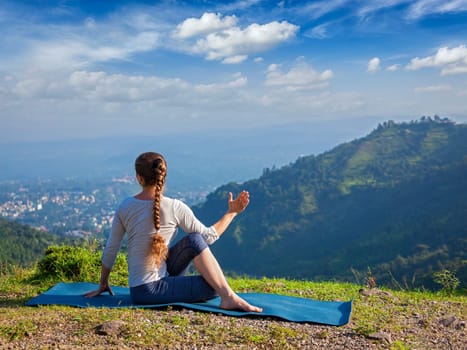 Hatha yoga outdoors - sporty fit woman doing yoga asana Parivrtta Marichyasana (or ardha matsyendrasana) - seated spinal twist outdoors in mountains in the morning