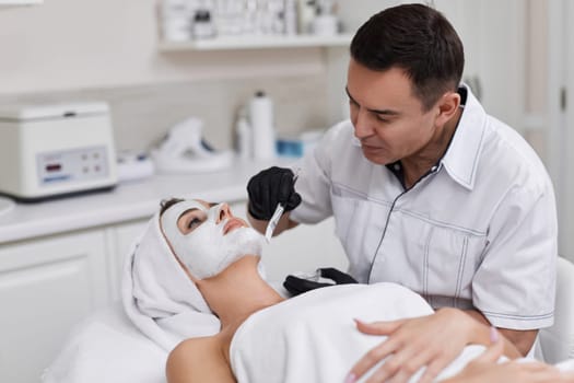 male beautician applies the mask to the face of beautiful woman in the spa salon. facial skin care concept.