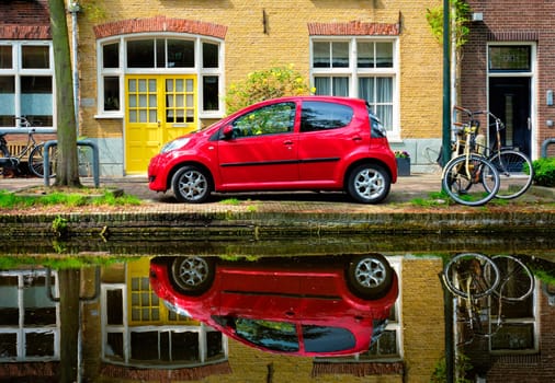 Red car on canal embankment in street of Delft with reflection and bicycle. Delft, Netherlands