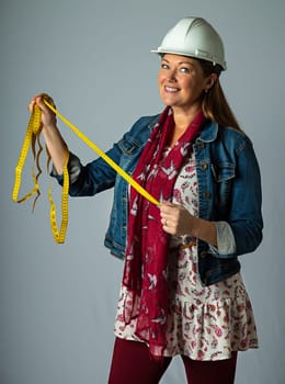 Forty year old woman, wearing bohemian style clothing and a hard hat, holding a yellow measuring tape, isolated on a white background