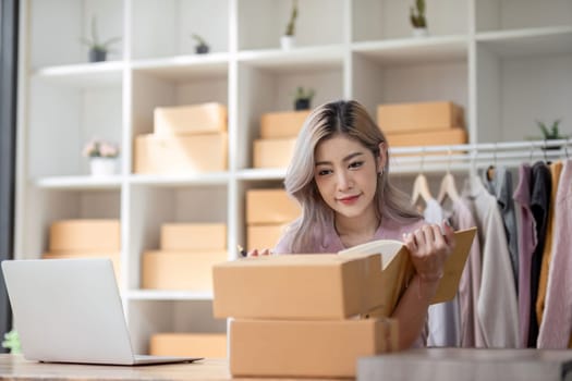 Female entrepreneur writes on parcel box after reading address on notebook. Online seller concept.