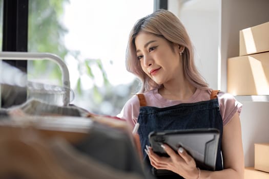 businesswoman owner an online trading business is using tablet to check the stock of the clothes on the rail rack at small office at home. work from home, online delivery, SME..