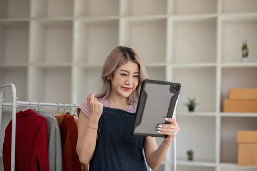 businesswoman owner an online trading business is using tablet to check the stock of the clothes on the rail rack at small office at home. work from home, online delivery, SME..
