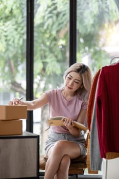 Female entrepreneur writes on parcel box after reading address on notebook. Online seller concept.