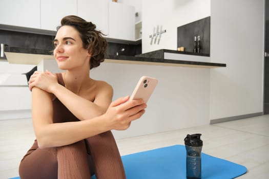 Portrait of sporty girl with mobile phone, sitting on yoga mat, using smartphone during workout at home, watching fitness turorial on telephone. Sport and wellbeing concept