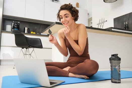 Portrait of young female athlete, yoga girl paying for online classes, purchasing remote training with gym instructor, sitting on her rubber mat with laptop and credit card.