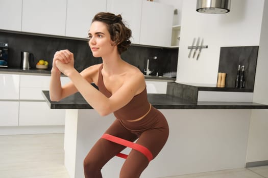 Healthy lifestyle and wellbeing. Young woman with sport equipment in her kitchen, doing squats with rubber elastic band, exercising, workout at home.