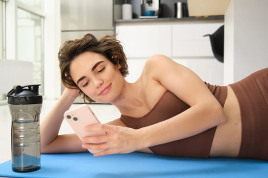 Online fitness and wellbeing. Portrait of young woman lying on yoga mat, looking at smartphone app, looking at workout tips, doing training at home in activewear.