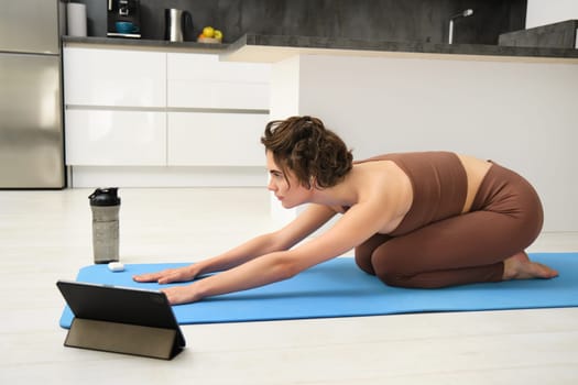 Image of young sportswoman does yoga at home, watches gym instructor tutorial video on tablet, stretches body, fitness training exercises, pilates class online from home.