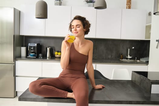 Happy brunette sportswoman, girl in activewear drinking glass of orange juice, sitting in her kitchen counter and smiling.