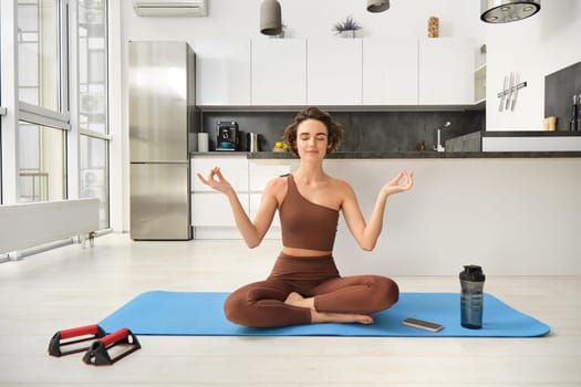 Smiling girl athlete, sportswoman doing yoga at home in activewear, sitting on yoga rubber mat in lotus pose, meditating, practice mindfulness exercises.